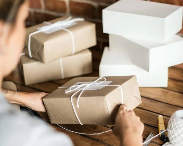 Hands wrapping a gift box with brown paper and white string on a wooden table.
